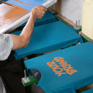 Artisan applying bright orange screen print onto a blue t-shirt using a squeegee, showcasing large-volume promotional apparel.
